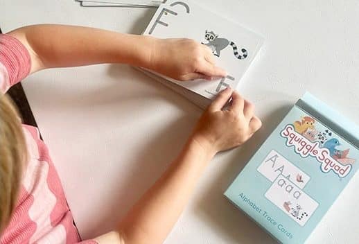 Child tracing an alphabet card featuring a lemur illustration from a set labeled "Squiggle Squad Alphabet Trace Cards.