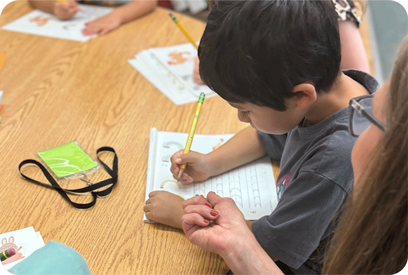 A young child writes in a workbook with a pencil while an adult guides their hand at a classroom table.