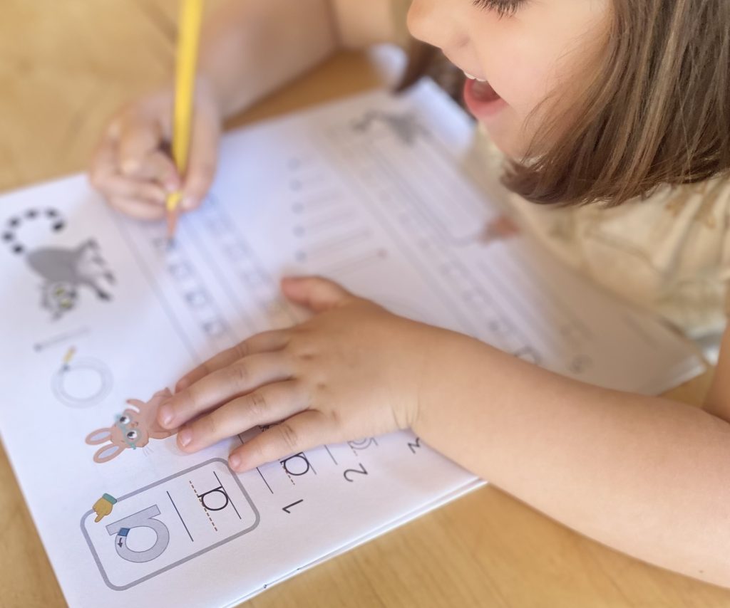 A child writes with a pencil on a worksheet featuring numbers and animal illustrations at a wooden table.