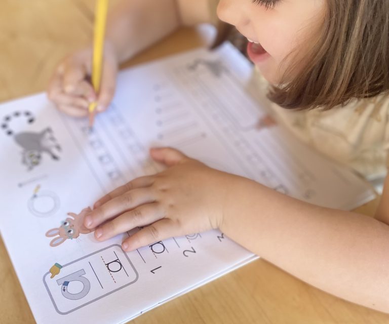 A child writes with a pencil on a worksheet featuring numbers and animal illustrations at a wooden table.